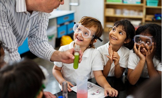 Three child students wearing safety goggles looking at their teacher holding a glass beaker