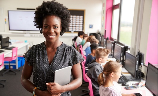 A teacher holding a notebook standing in front of her class working on computers