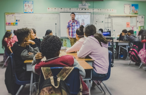 students sitting in desks in a classroom with a teacher standing in front