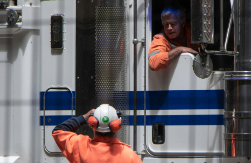 Photo of a man sitting inside of an eighteen-wheeler truck talking to a man standing next to the truck. Both men are wearing neon orange work jackets and the man standing outside of the truck is wearing a hard hat.