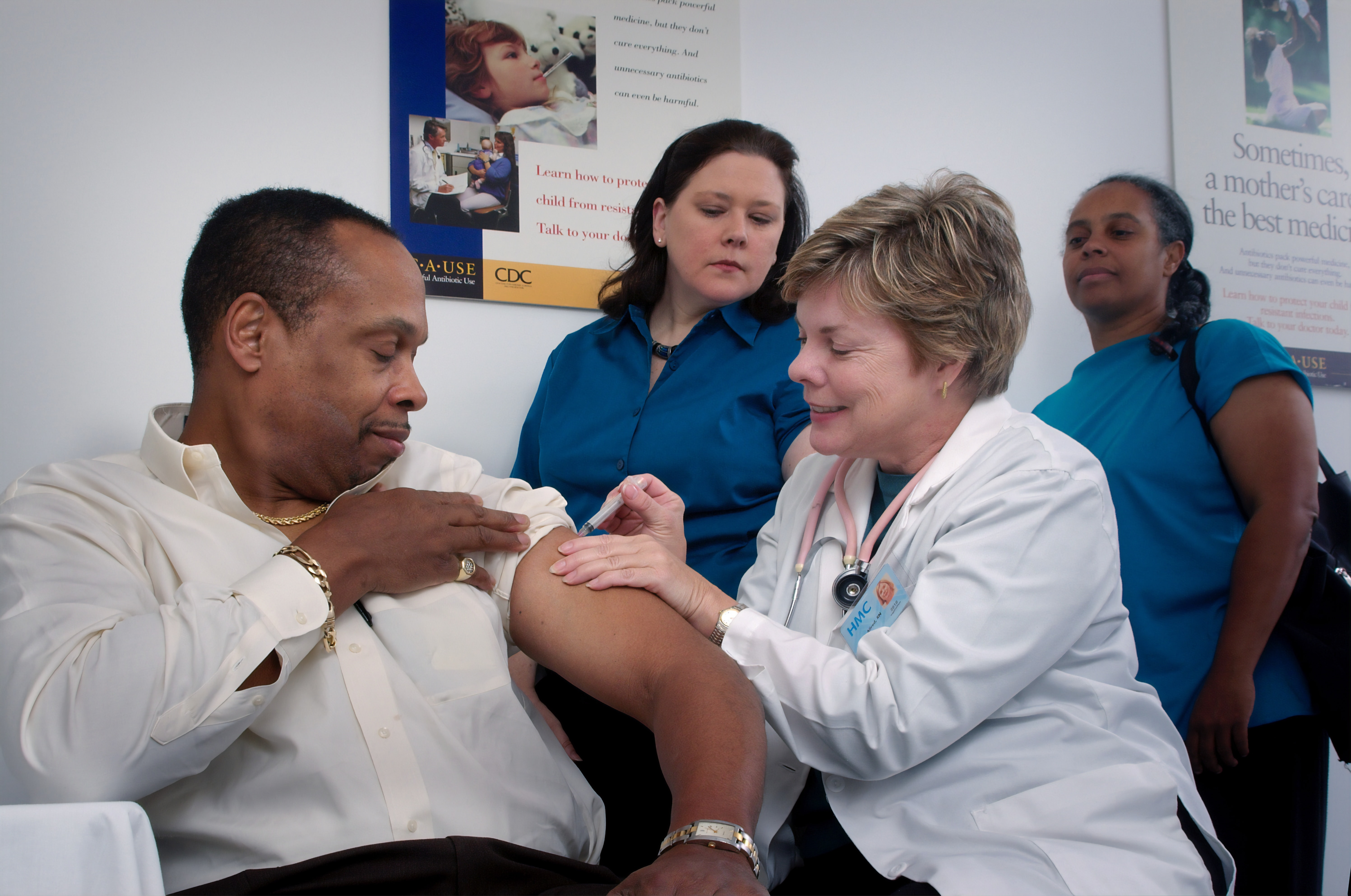 A man wearing a white shirt receives a vaccination by a doctor as two nurses look on. 