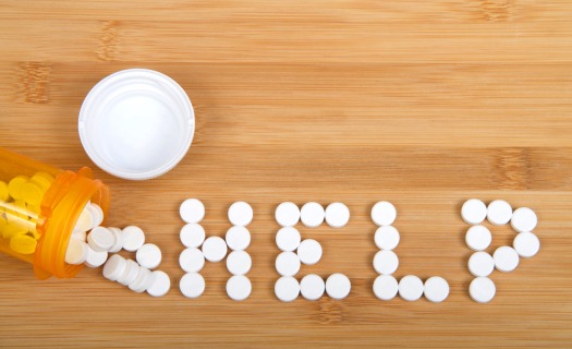 The word HELP spelled out on a wood table in white pills. There is a prescription bottle open and lying on its side.