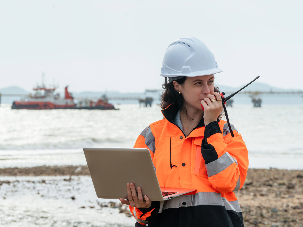Woman in PPE speaks into radio while a tugboat floats behind her