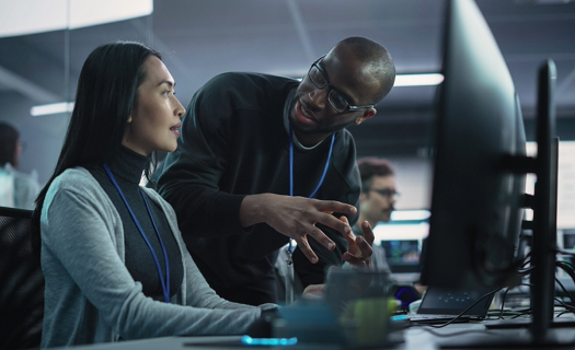 Colleagues working in an in-person tech environment, chatting in front of a desktop.