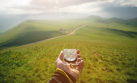 Person holding a compass out in front of them while walking across a ridge top in a rural area. The horizon is foggy.