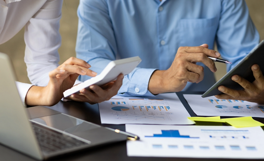 Two people in business attire review charts and graphs while making calculations on a tablet and a calculator.
